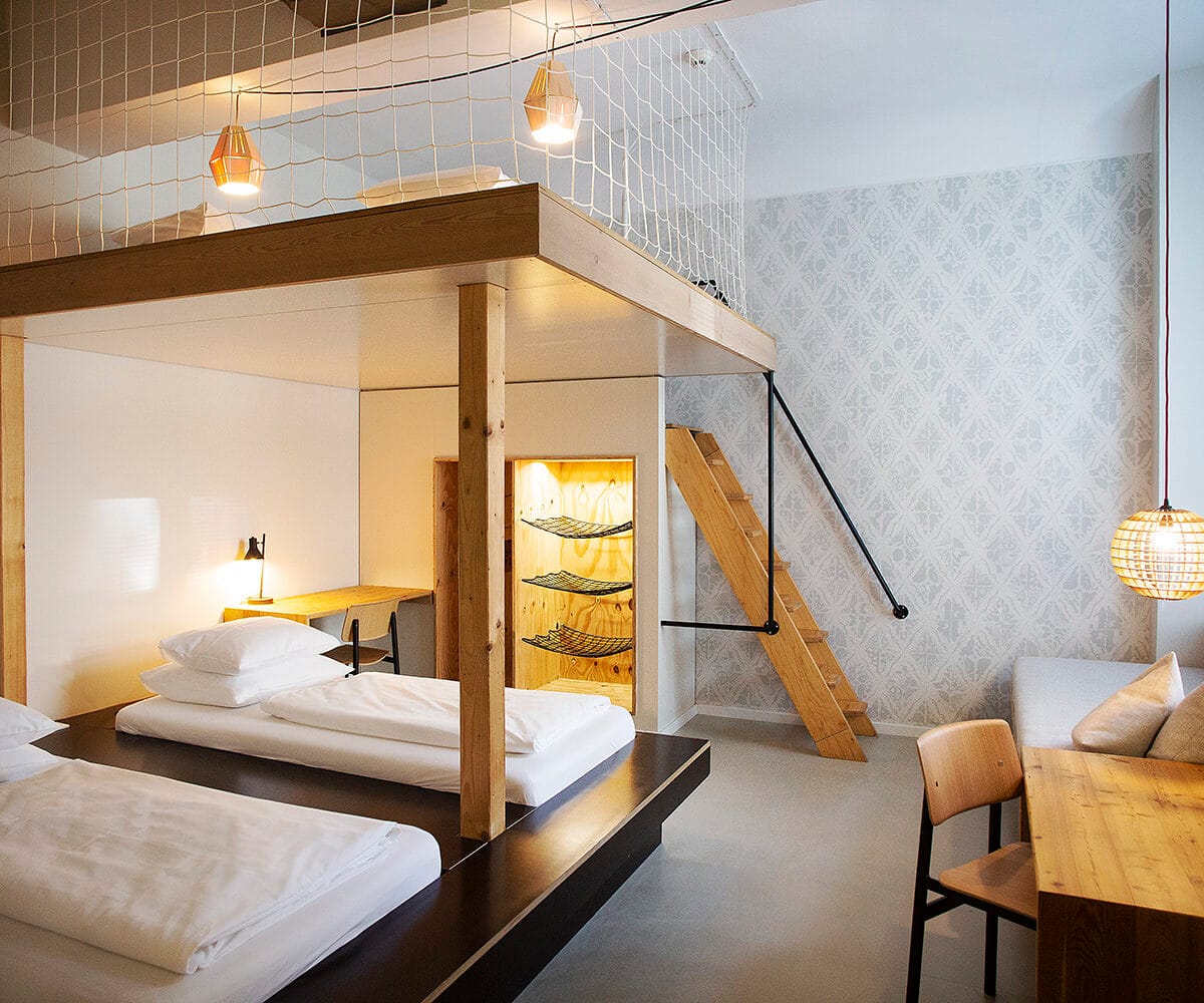 Modernes Zimmer mit zwei Betten, Holzregalen, einer Treppe und einem gemütlichen Sitzbereich. Helle, stilvolle Einrichtung.