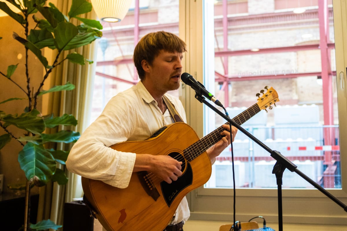 Mann mit Gitarre singt vor einem Mikrofon, umgeben von Pflanzen und großen Fenstern.